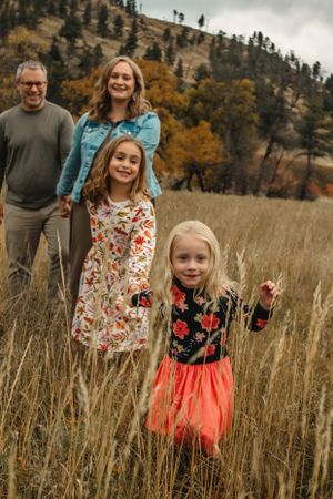 A happy family stands together in a field of tall grass with autumn mountains and trees in the background.