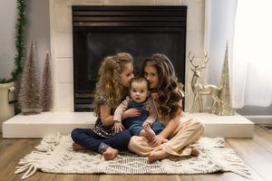 Family sitting together on a cozy white rug in front of a fireplace with decorative deer and holiday trees.