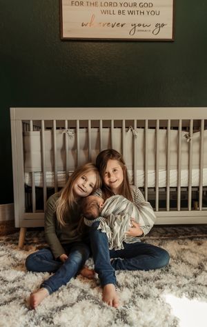 Siblings sit together on a plush rug in front of a white crib beneath an inspirational wall sign.