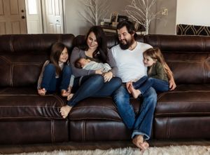 Family relaxes together on a brown leather couch in a cozy living room with a decorative white tree in background.