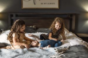 Three siblings share a cozy moment together while lounging on a gray bed beneath a rustic wooden headboard.