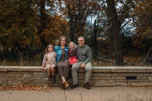 A family of four sits together on a stone wall surrounded by autumn trees with fallen leaves scattered around.