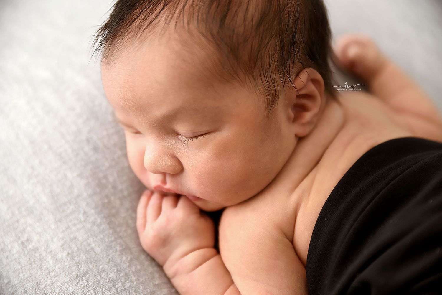 Close-up of a sleeping newborn baby with hands tucked under chin on gray fabric.