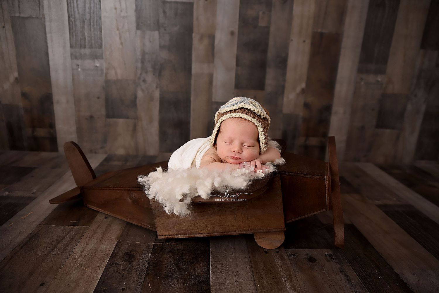 Newborn baby sleeping in a wooden toy airplane prop wearing a cream knit aviator bonnet hat.