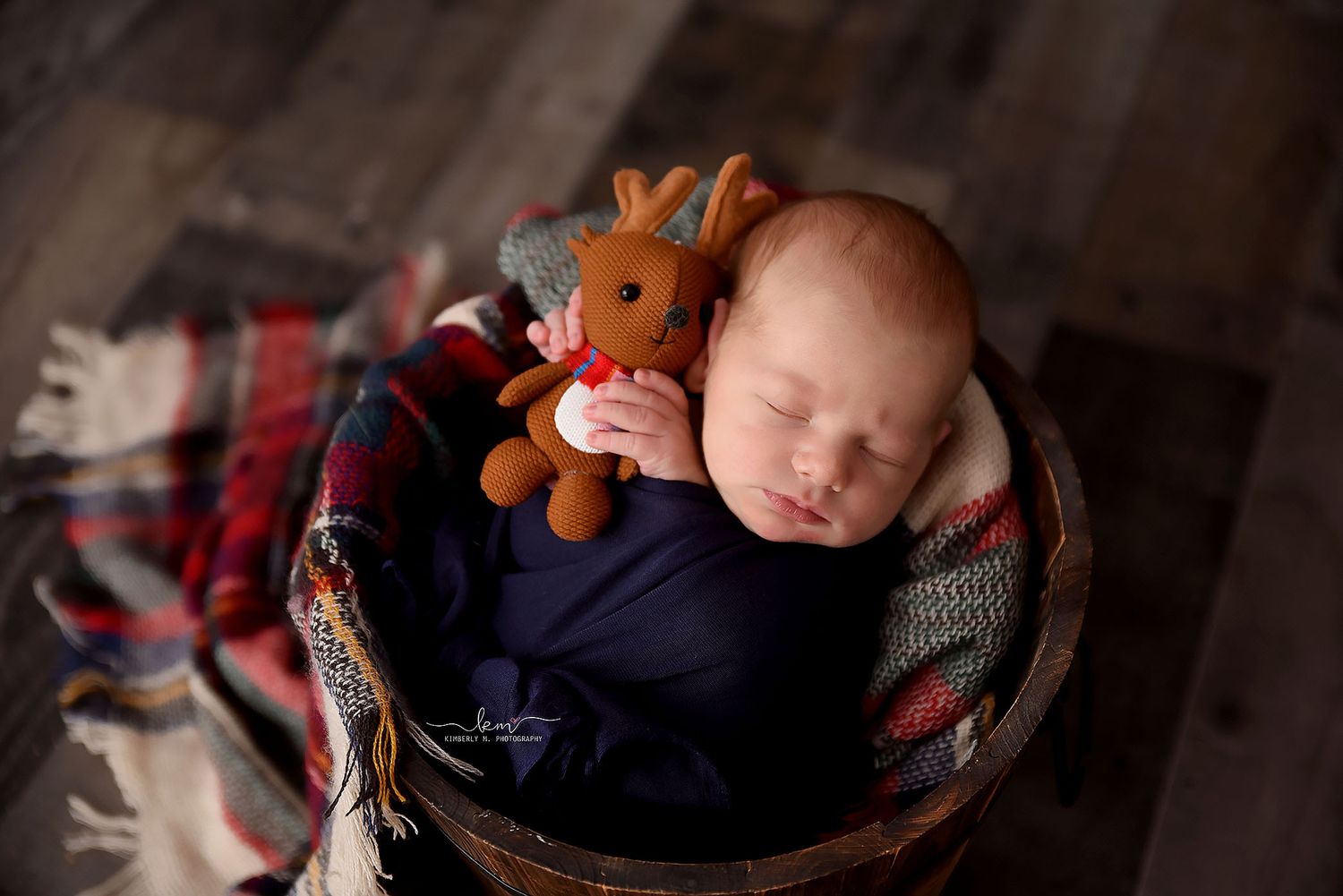 Sleeping newborn baby boy wrapped in navy blue swaddle holding a small reindeer stuffed animal in a basket.
