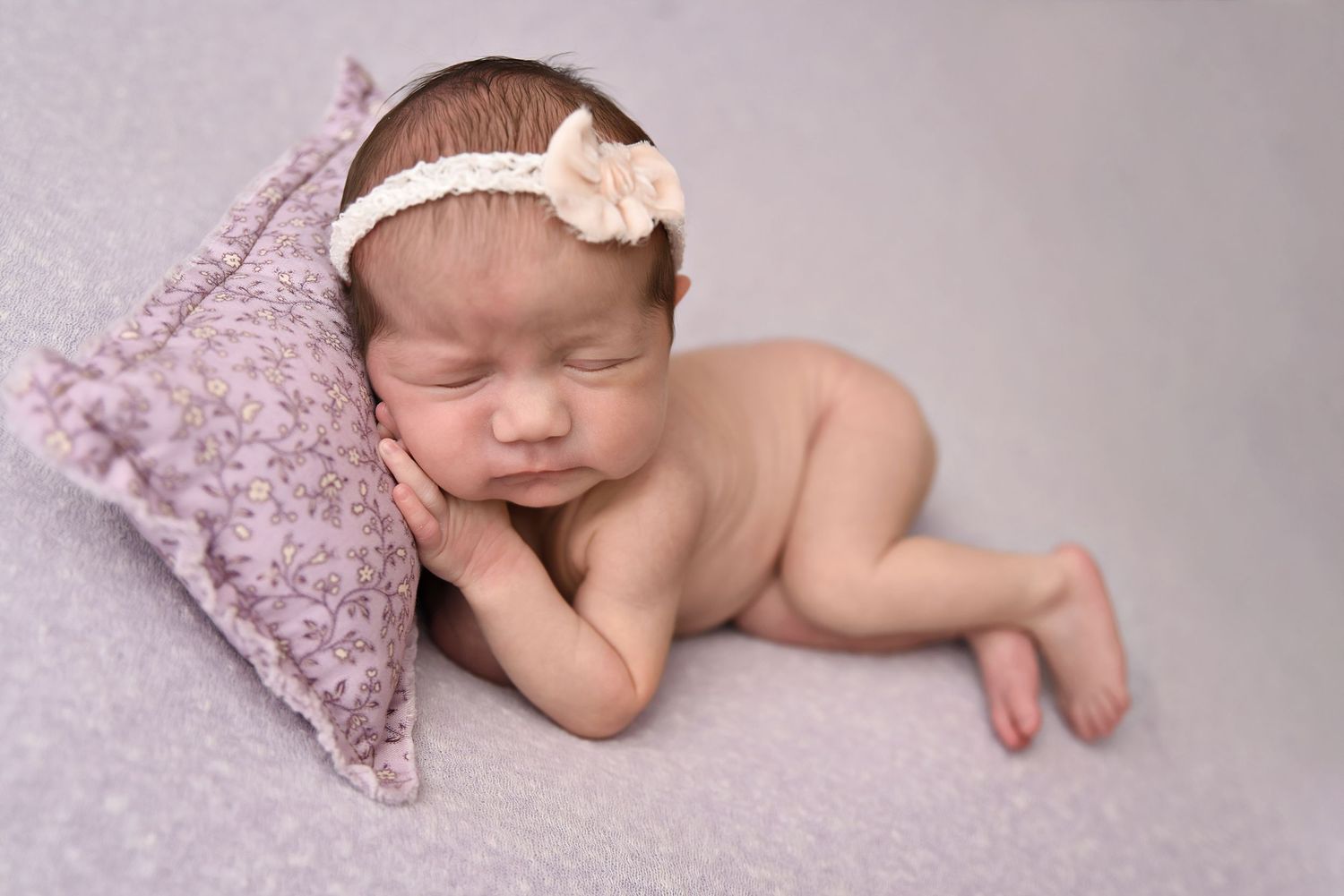 Sleeping newborn girl with floral headband resting on a small lavender pillow on a purple background.