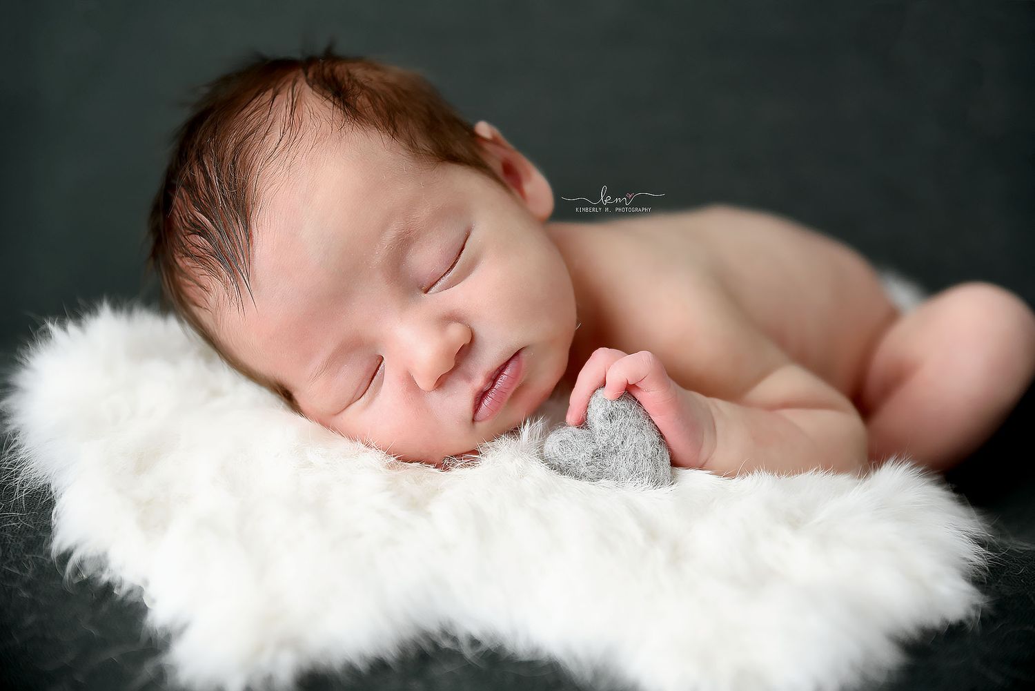 Newborn baby sleeping on white fur rug holding a small gray heart prop.