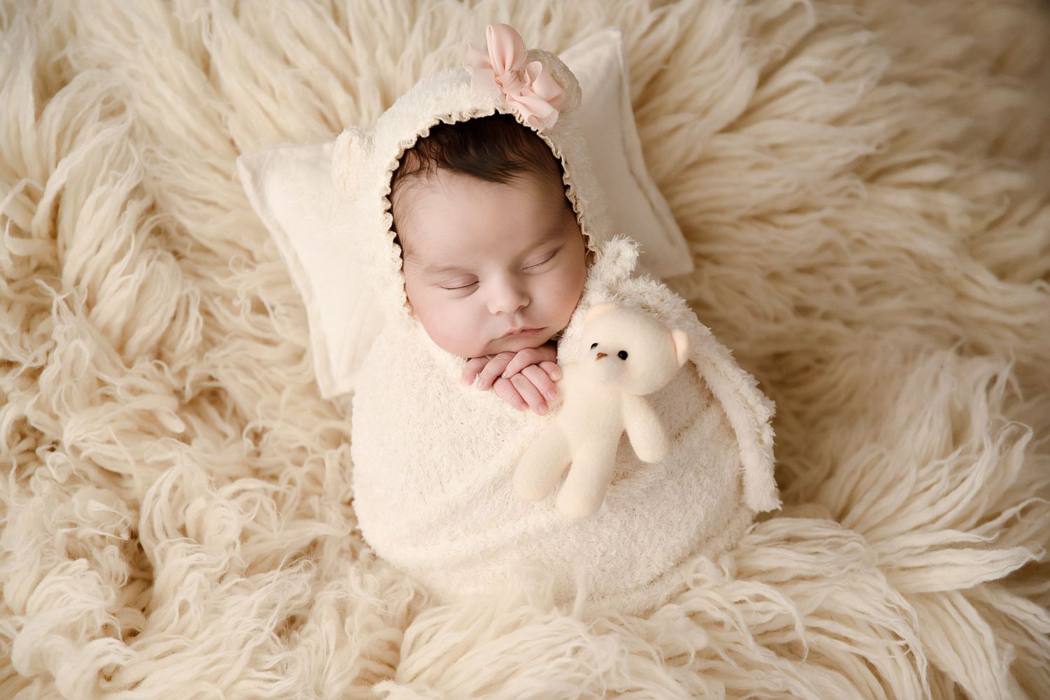 Sleeping newborn girl swaddled in cream wrap holding a stuffed bunny toy on a fluffy white fur blanket.