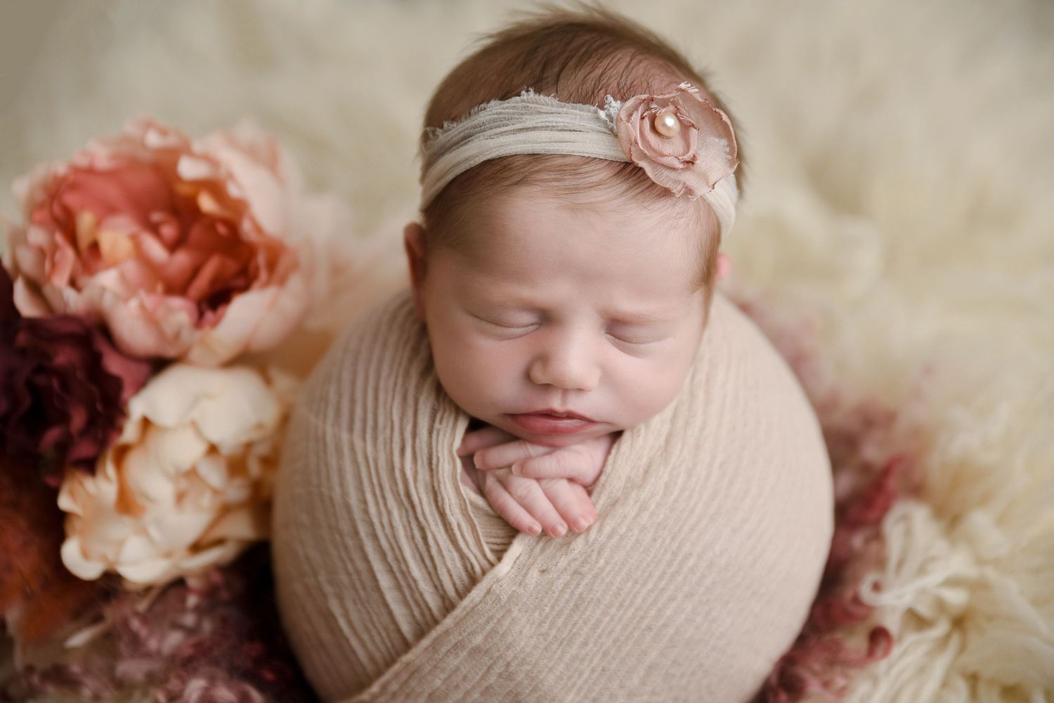 Newborn baby girl wrapped in a beige swaddle with a pink rose headband surrounded by blush flowers.