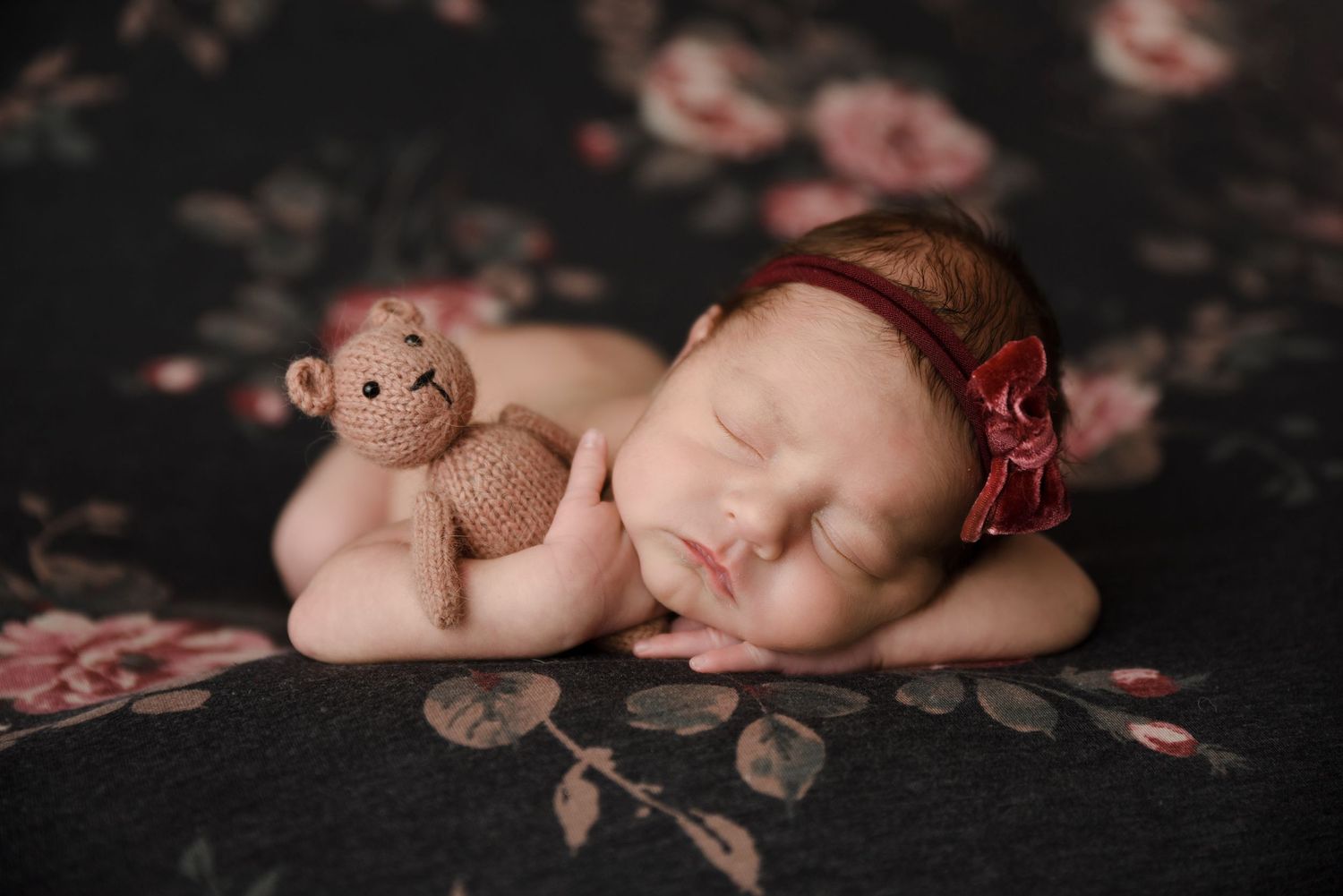 Sleeping newborn baby girl with a red floral headband cuddling a knit monkey toy on a floral backdrop.