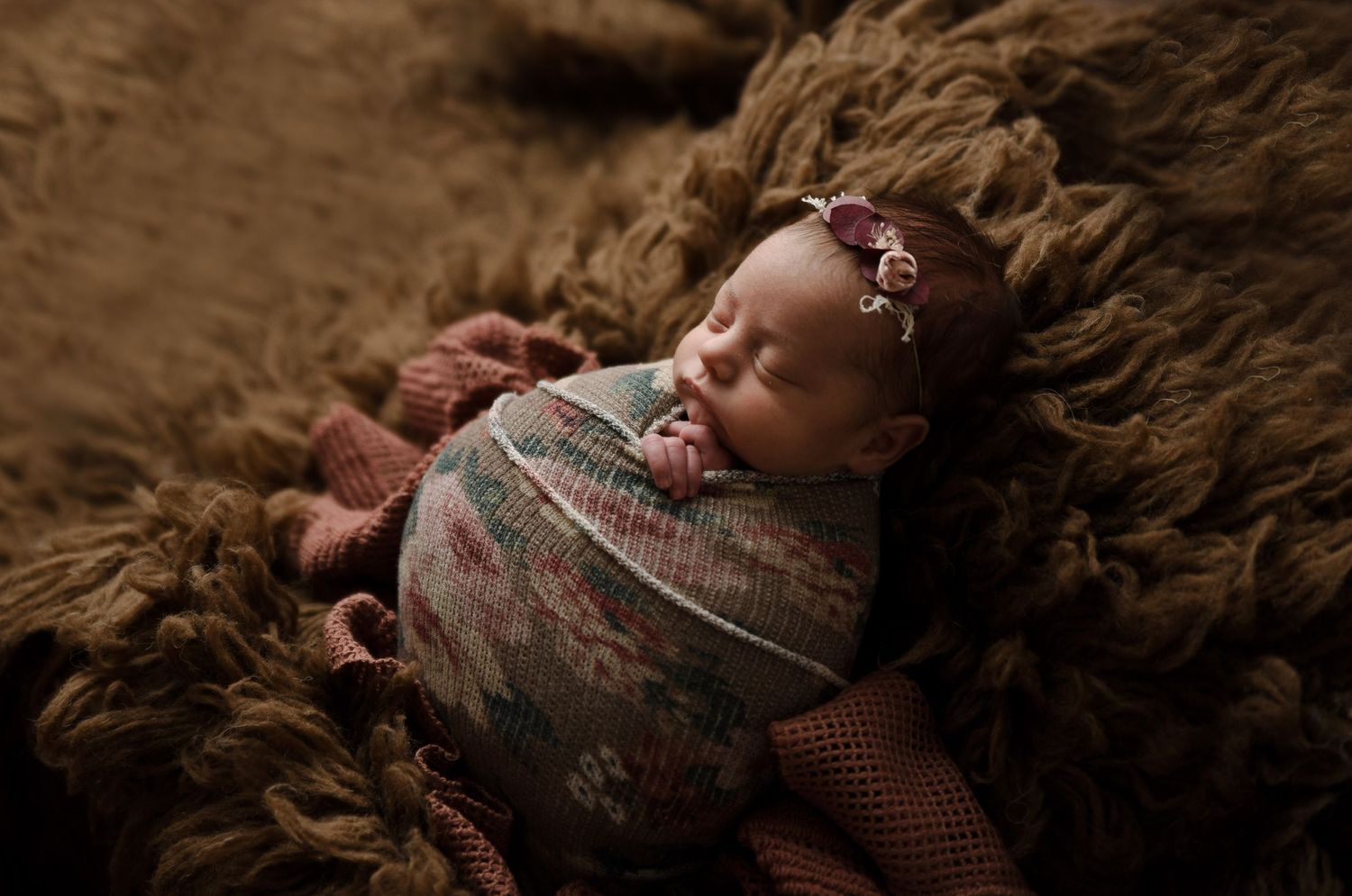 Newborn baby girl swaddled in a floral knit wrap with a flower headband, sleeping on a brown fluffy rug.