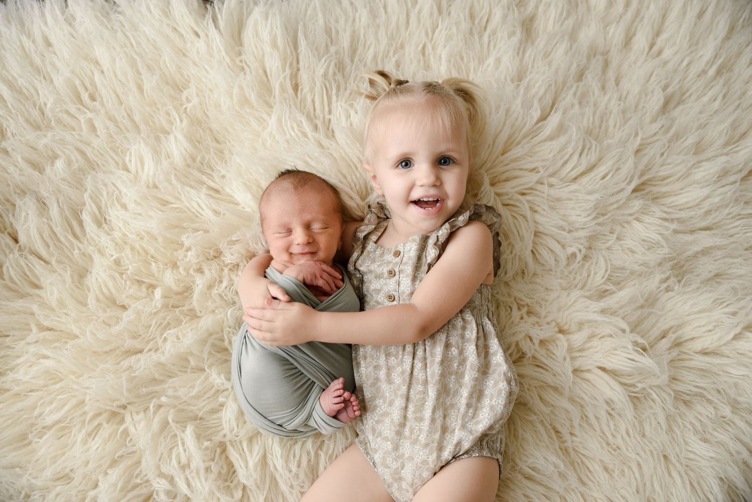 Smiling toddler girl in floral romper hugging swaddled newborn sibling on a cream fluffy rug.