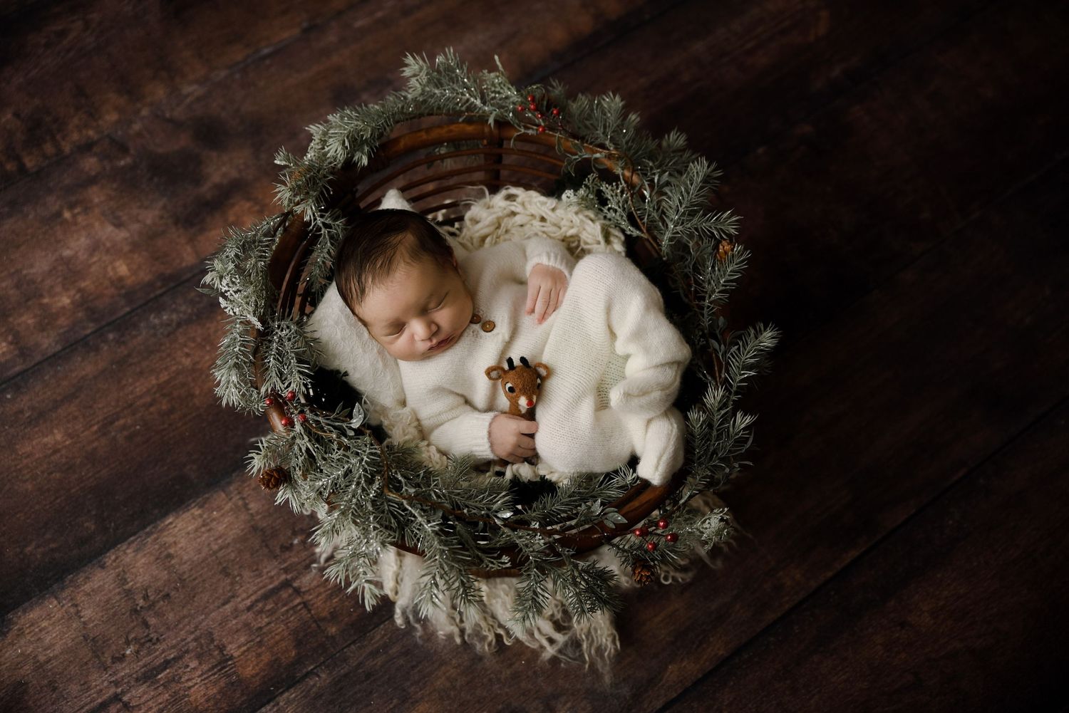 Sleeping newborn baby in a white outfit holding a toy, nestled in a wreath basket on dark wood floor.