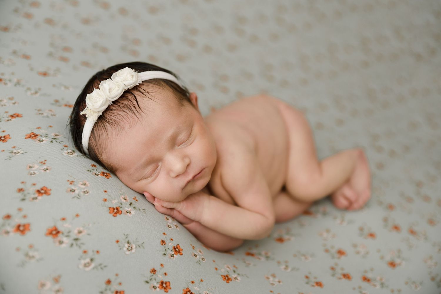 Peaceful sleeping newborn baby girl with white flower headband posed on a floral patterned backdrop.