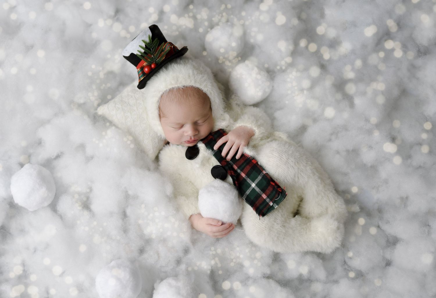 Newborn baby dressed in snowman outfit with top hat, sleeping on white pillow surrounded by snowballs.