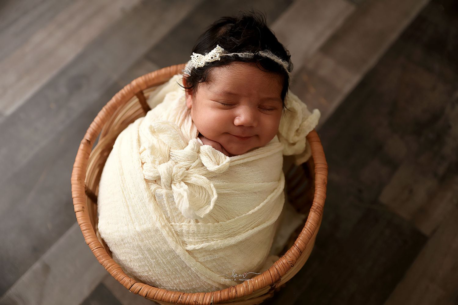 Sleeping newborn baby wrapped in cream swaddle with delicate headband posed inside a wicker basket.