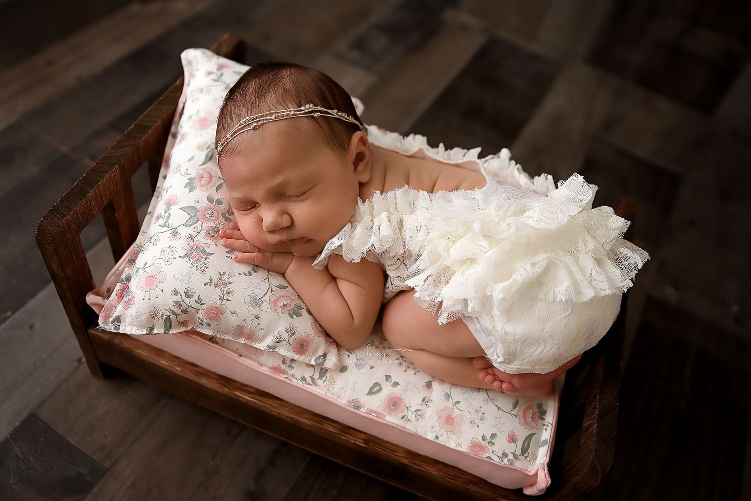 Newborn baby in white lace outfit sleeping on a floral pillow in a small wooden bed.