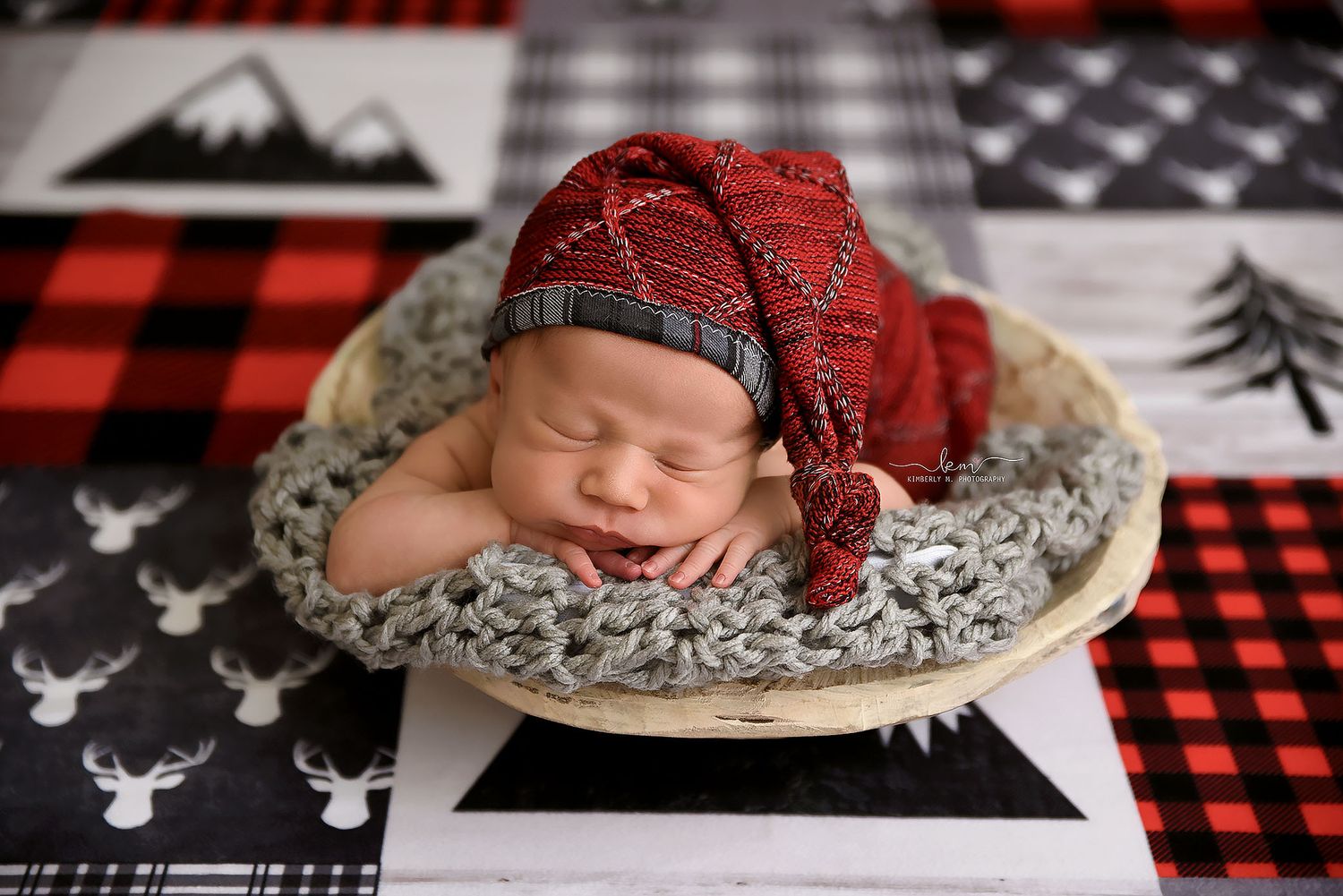 Sleeping newborn baby in red plaid wrap and matching nightcap posed in a wooden bowl with rustic holiday backdrop.