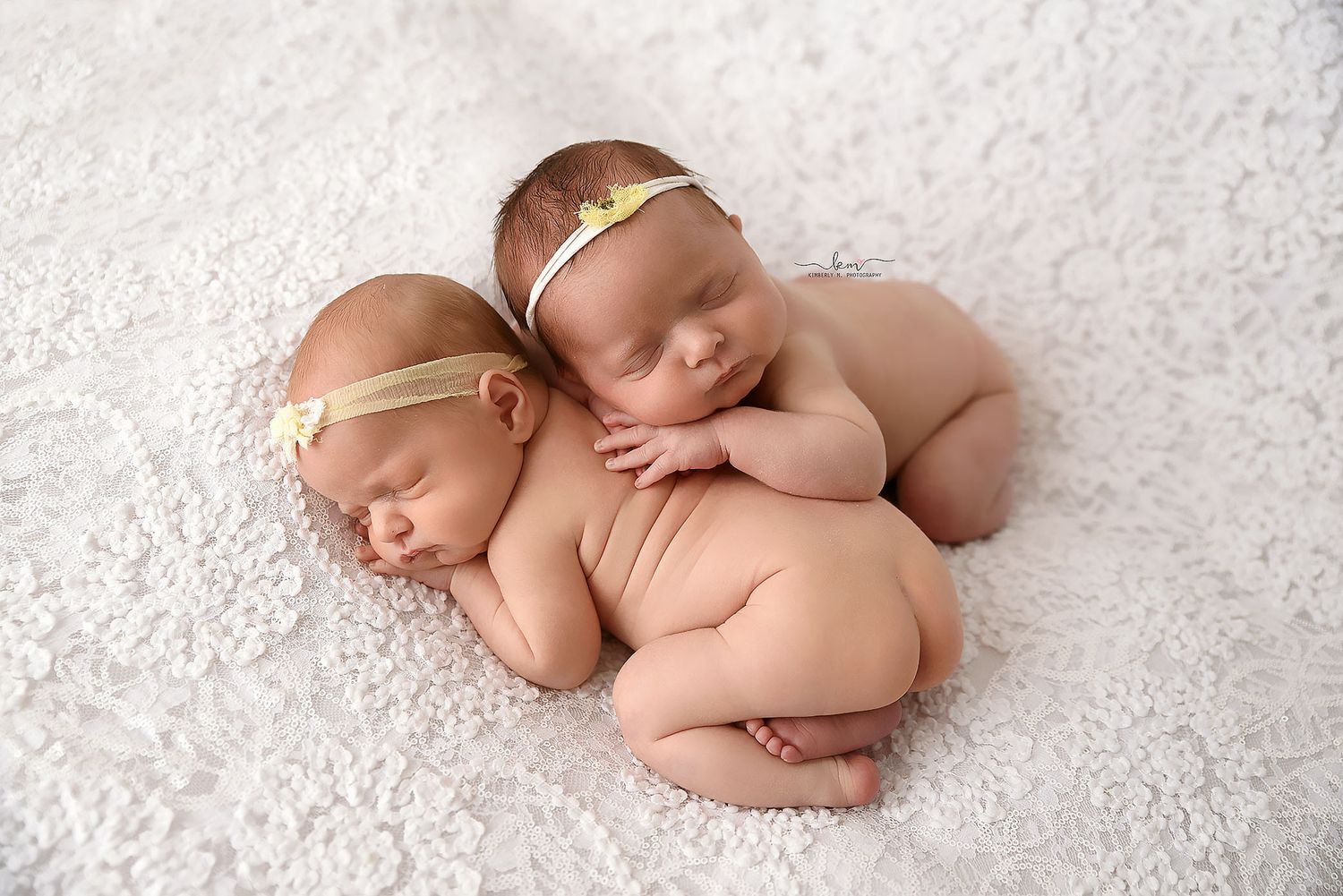 Twin newborn babies sleeping together on a white fluffy blanket with gold headbands.