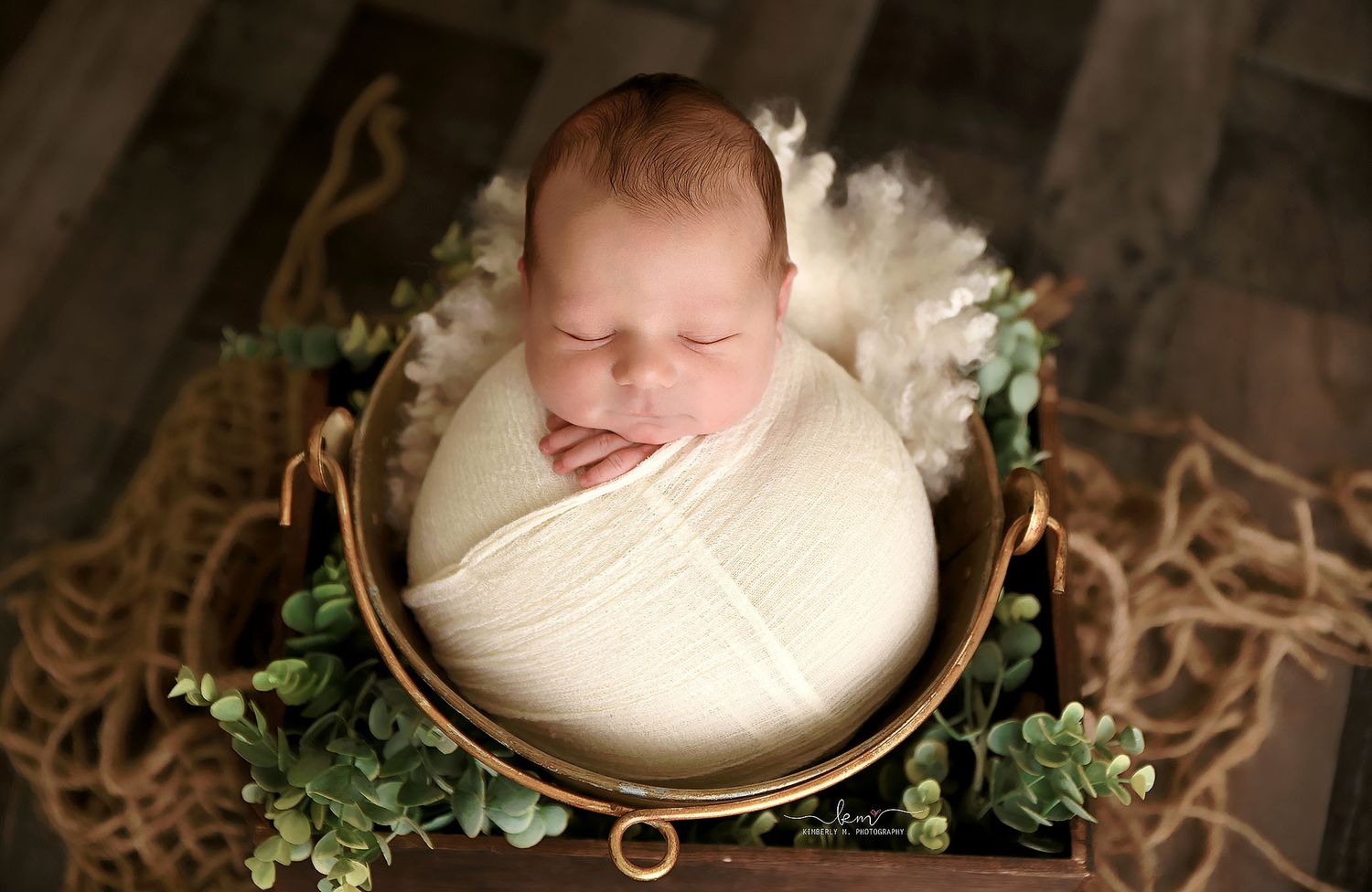 Sleeping newborn baby wrapped in white swaddle posed in a rustic metal bucket with green eucalyptus.