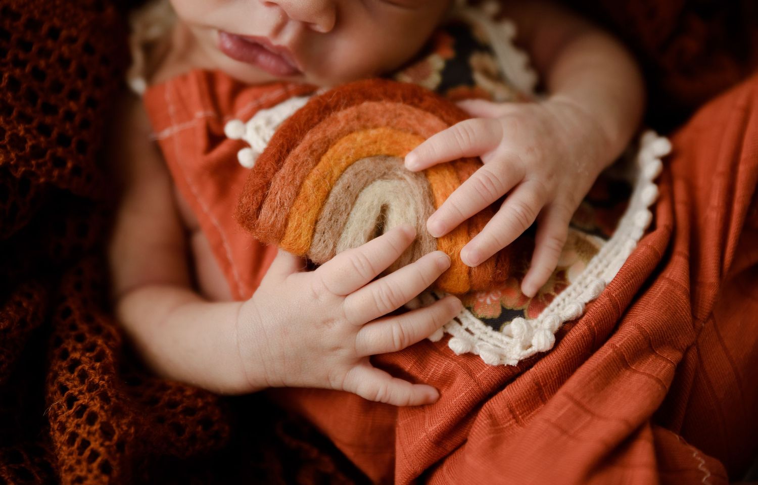 Close-up of baby in rust orange dress holding a handmade rainbow toy with crochet lace detail.