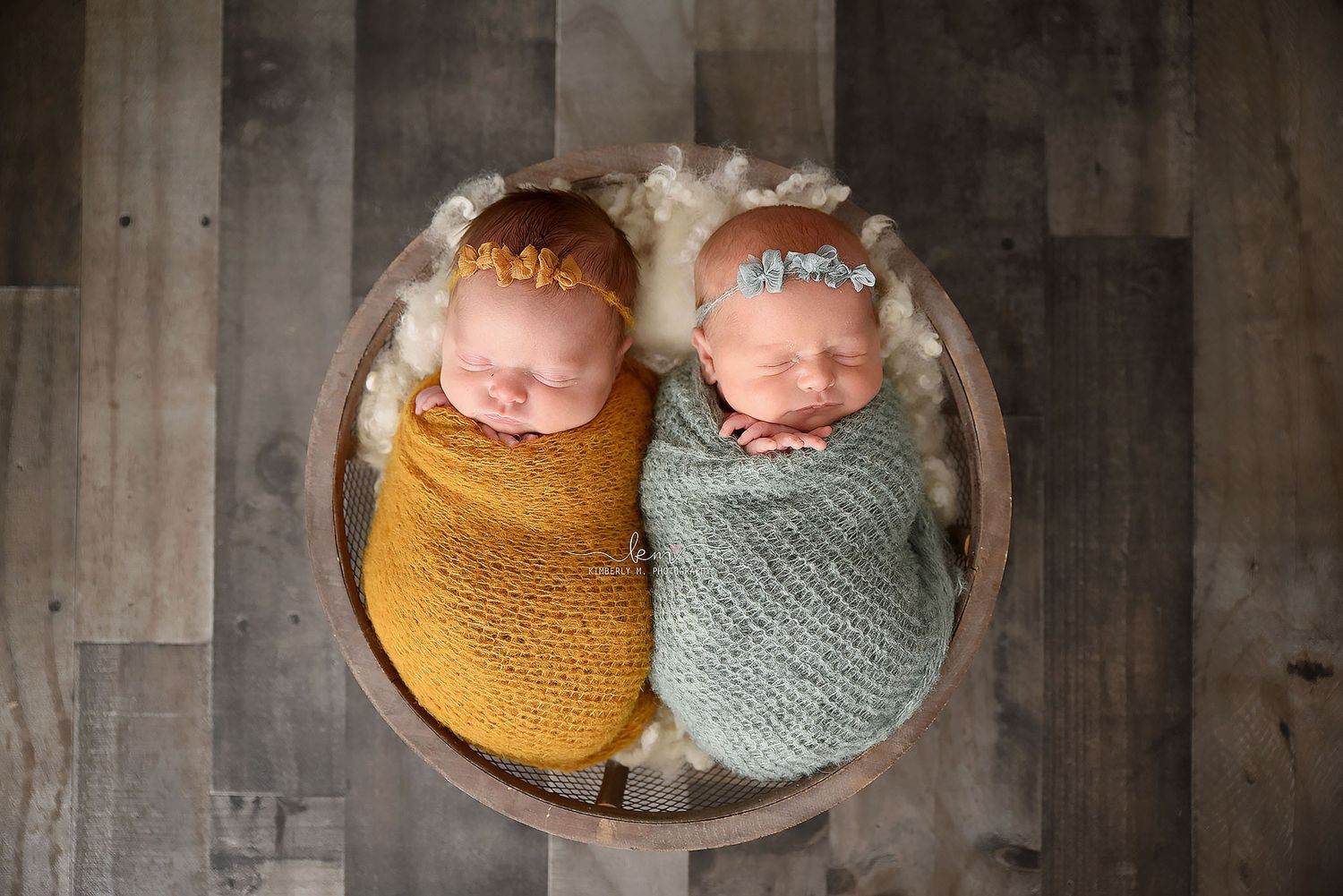 Two swaddled newborn twins sleeping in a wooden bowl, wrapped in gold and sage green knit blankets.