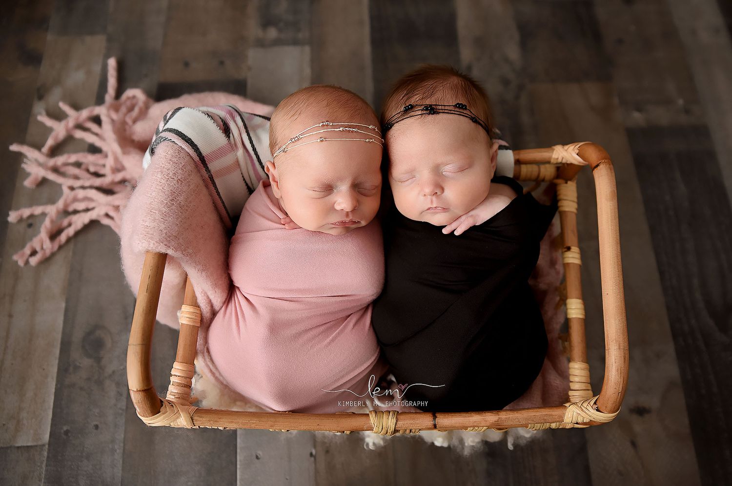 Two swaddled newborns sleeping side by side in a wicker basket, wrapped in pink and black knit blankets.