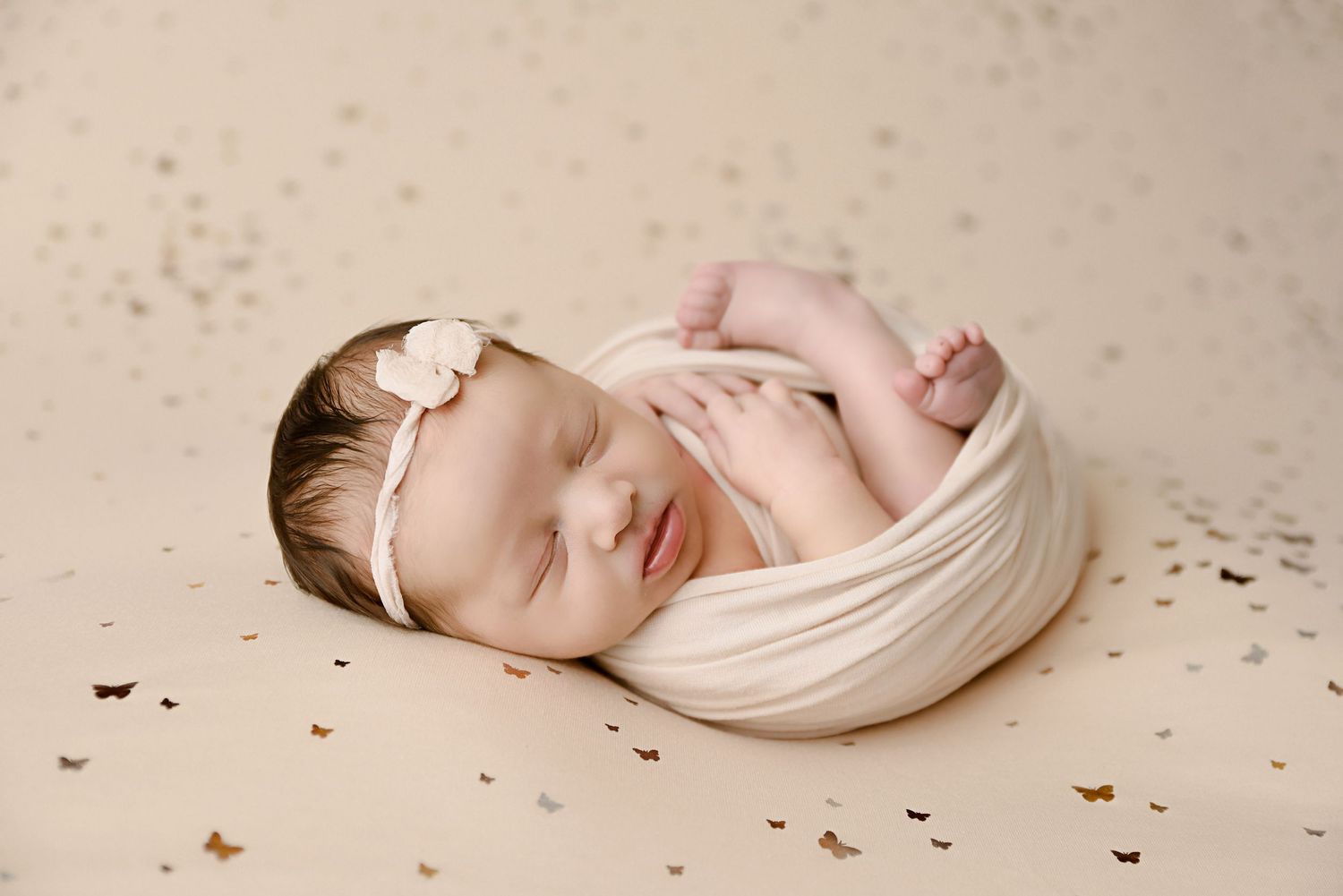 Sleeping newborn baby girl swaddled in cream wrap with bow headband on gold star-confetti backdrop.