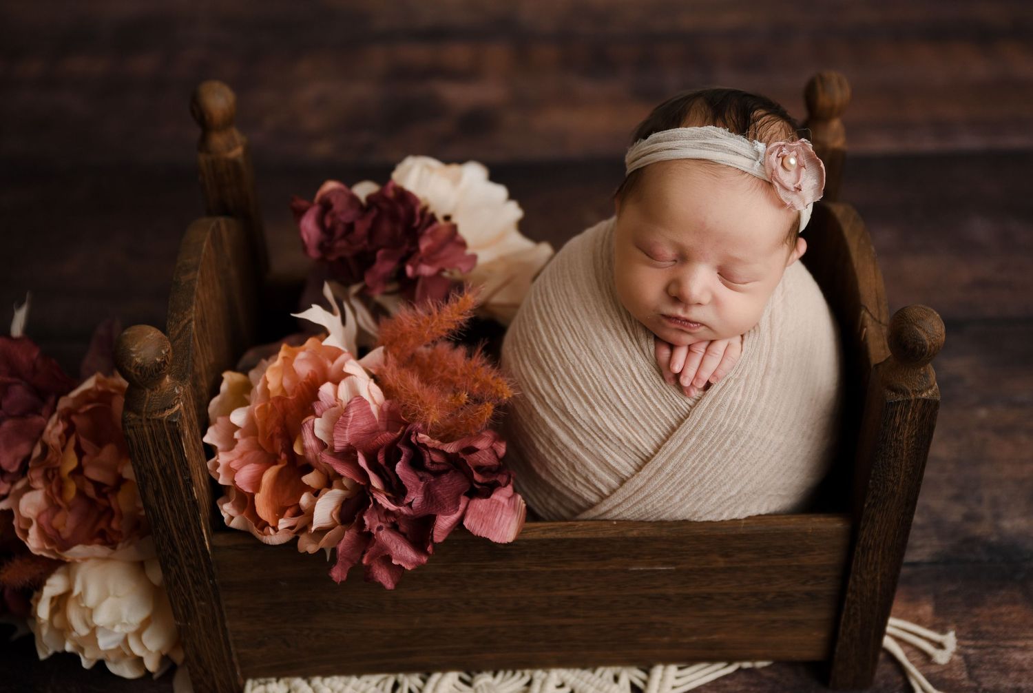 Swaddled newborn baby girl with floral headband sleeping in a rustic wooden bed with pink flowers.