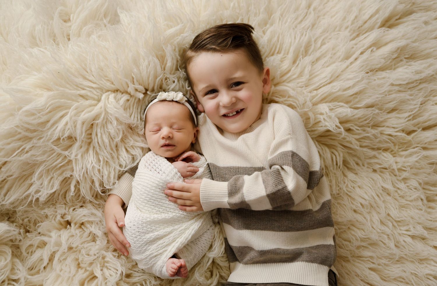 Smiling young boy lying on fluffy cream rug beside swaddled newborn baby sister with headband.