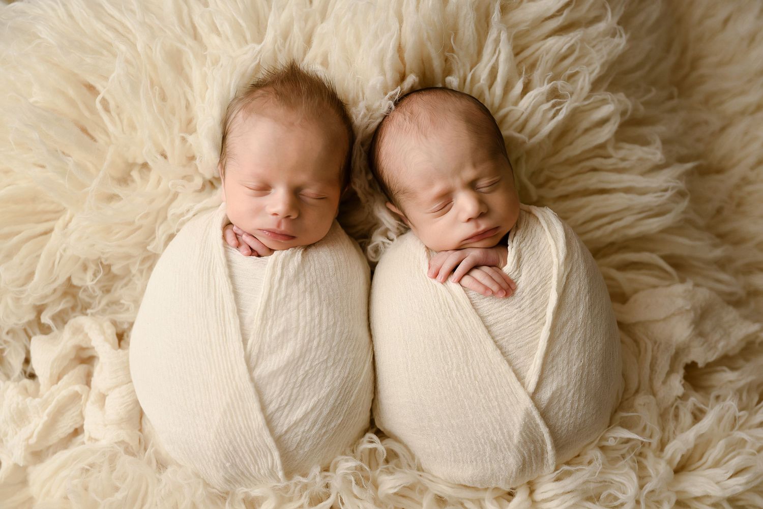 Two sleeping newborn twins swaddled in cream wraps lying side by side on a fluffy white fur blanket.
