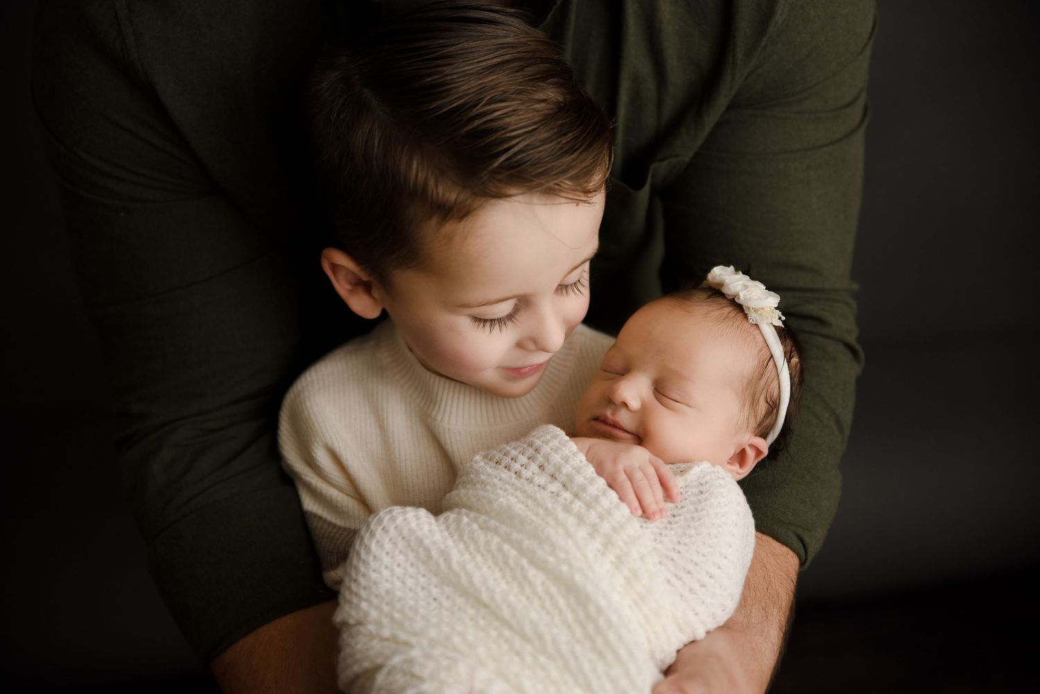 Young boy tenderly gazing at sleeping newborn baby sister wrapped in white, held by parent.