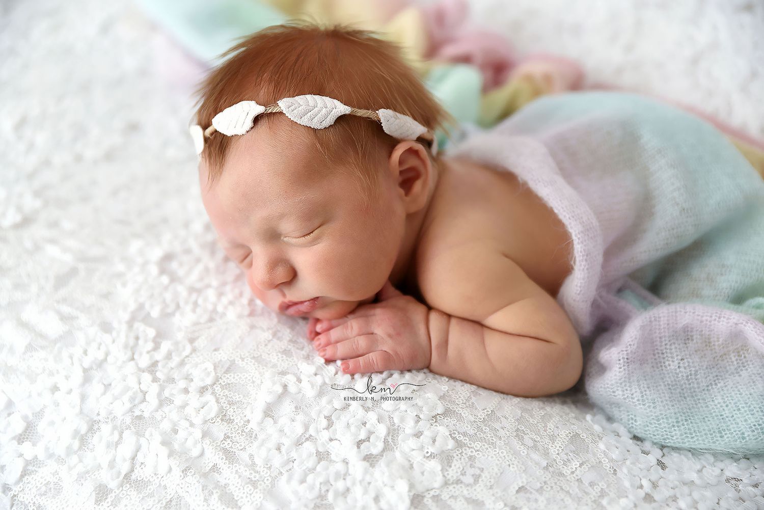 Peaceful sleeping newborn baby girl wearing a white leaf headband, resting on a soft white textured blanket.