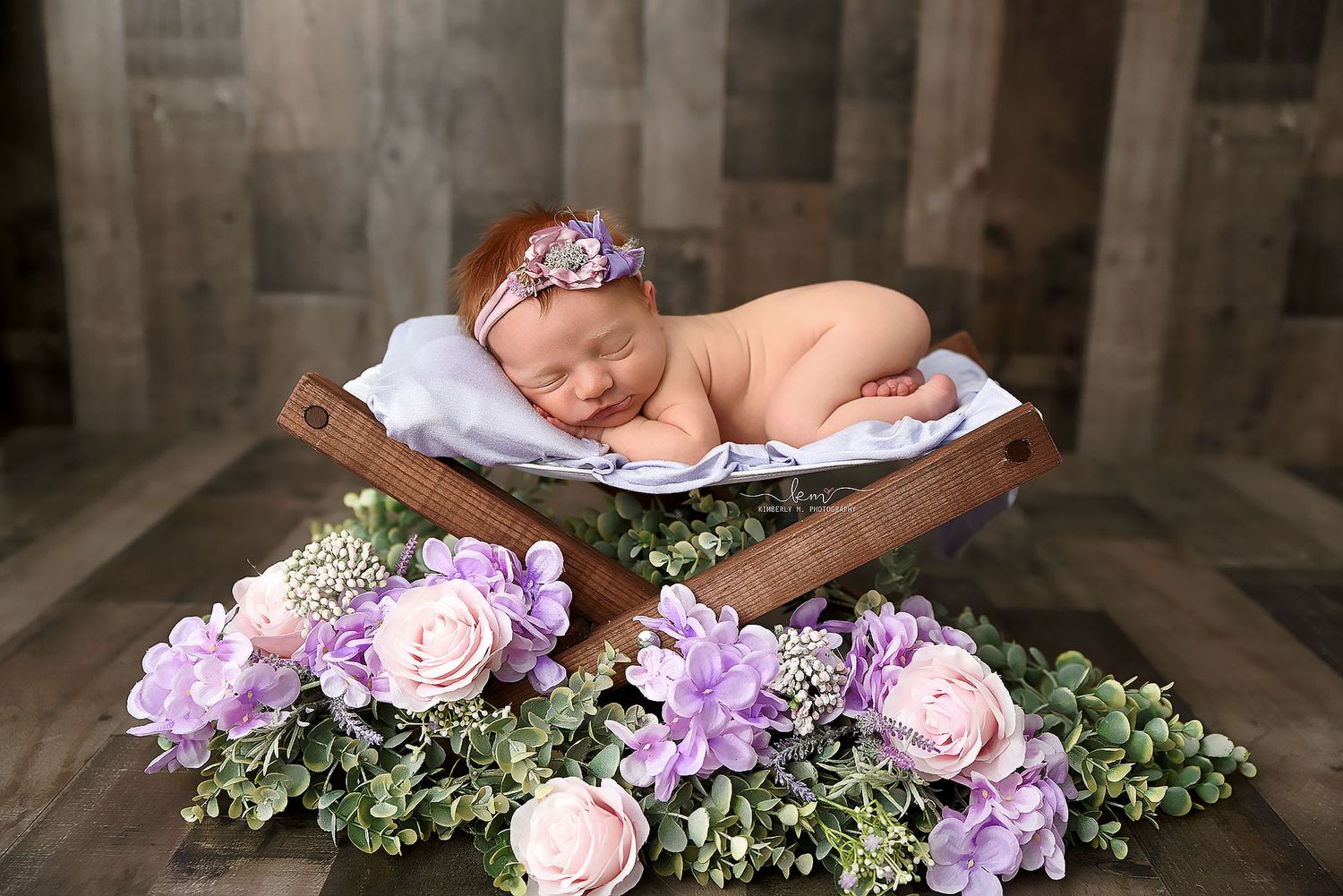 Sleeping newborn baby girl in a wooden bed prop surrounded by purple and pink flowers with a floral headband.