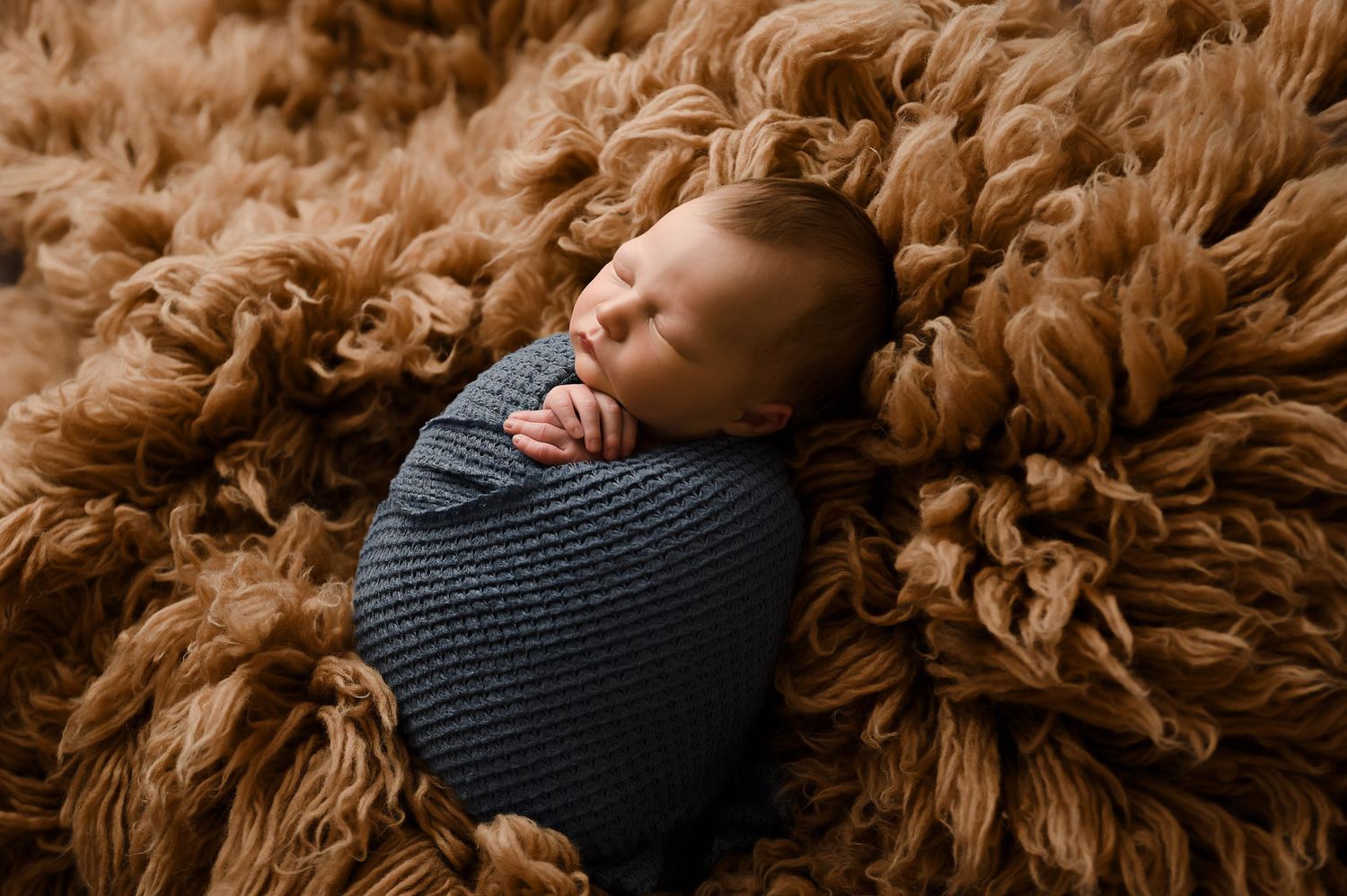 Peaceful newborn baby wrapped in grey knit swaddle sleeping on fluffy brown textured blanket.