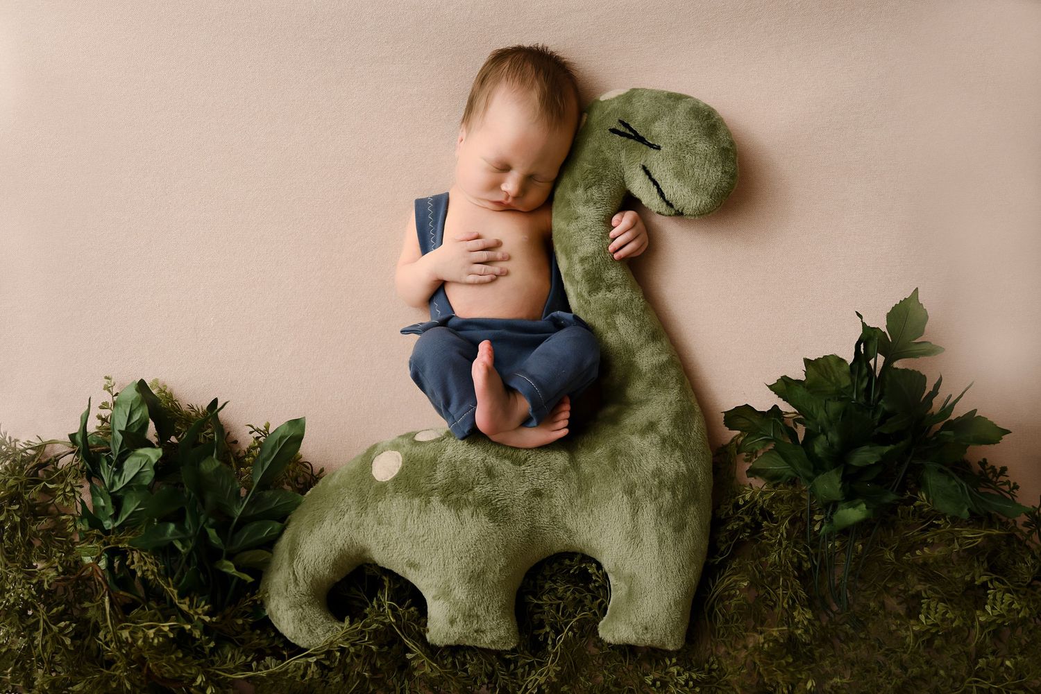 Sleeping newborn baby boy posed on green dinosaur prop surrounded by lush greenery on pink backdrop.