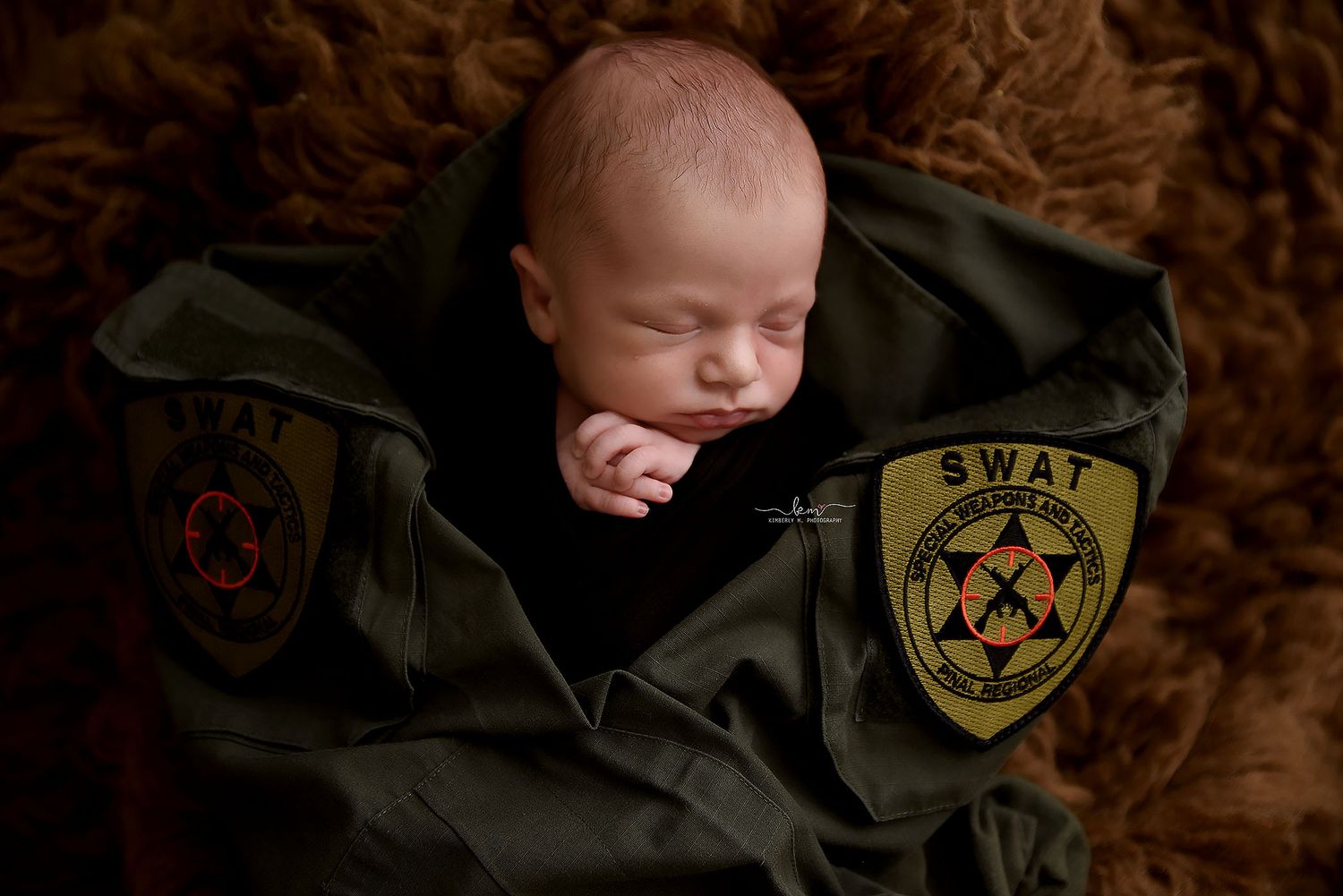 Sleeping newborn baby wrapped in a green SWAT uniform jacket on a brown fur blanket.