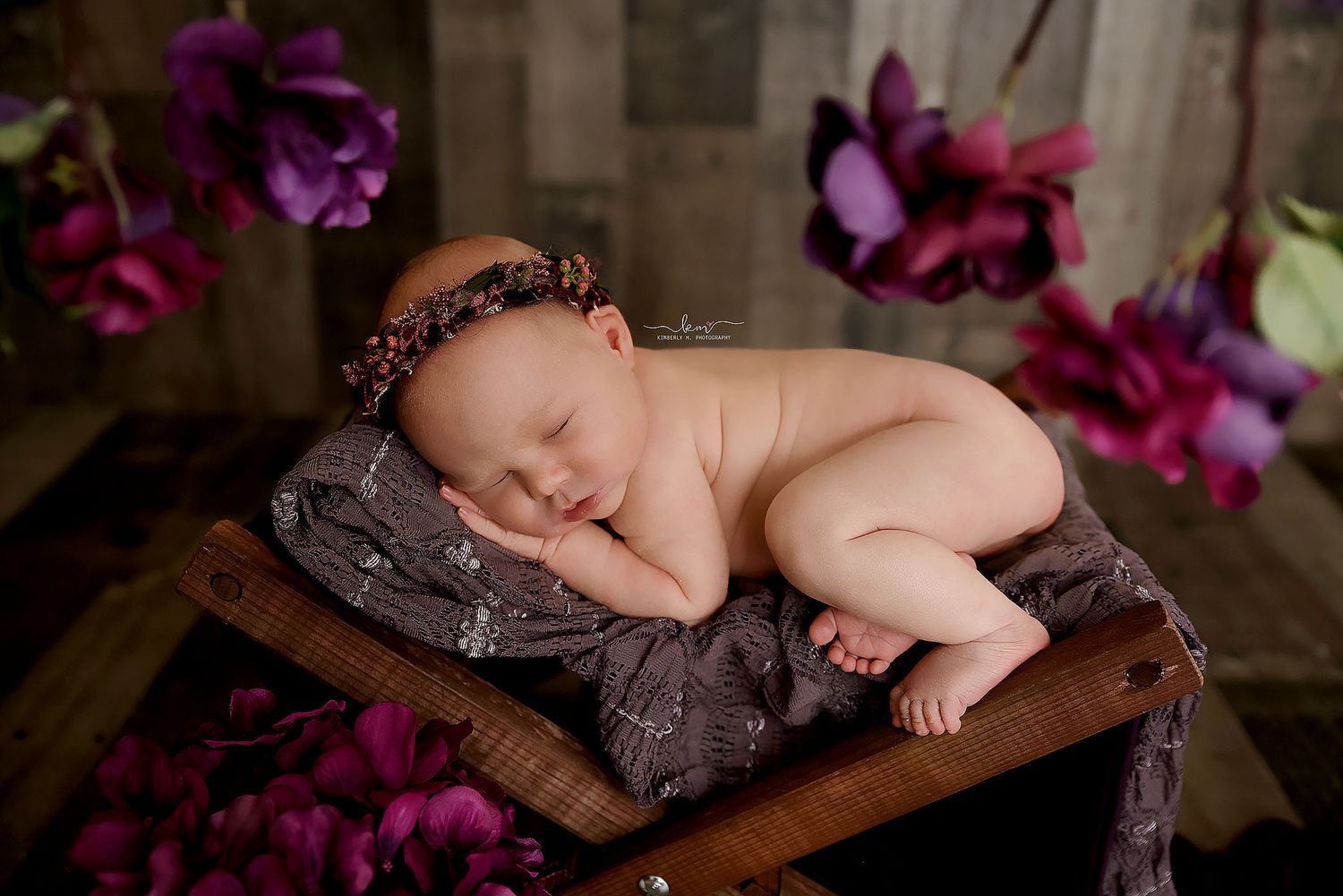 Peaceful newborn baby sleeping on purple fabric in a wooden crate surrounded by beautiful purple flowers.