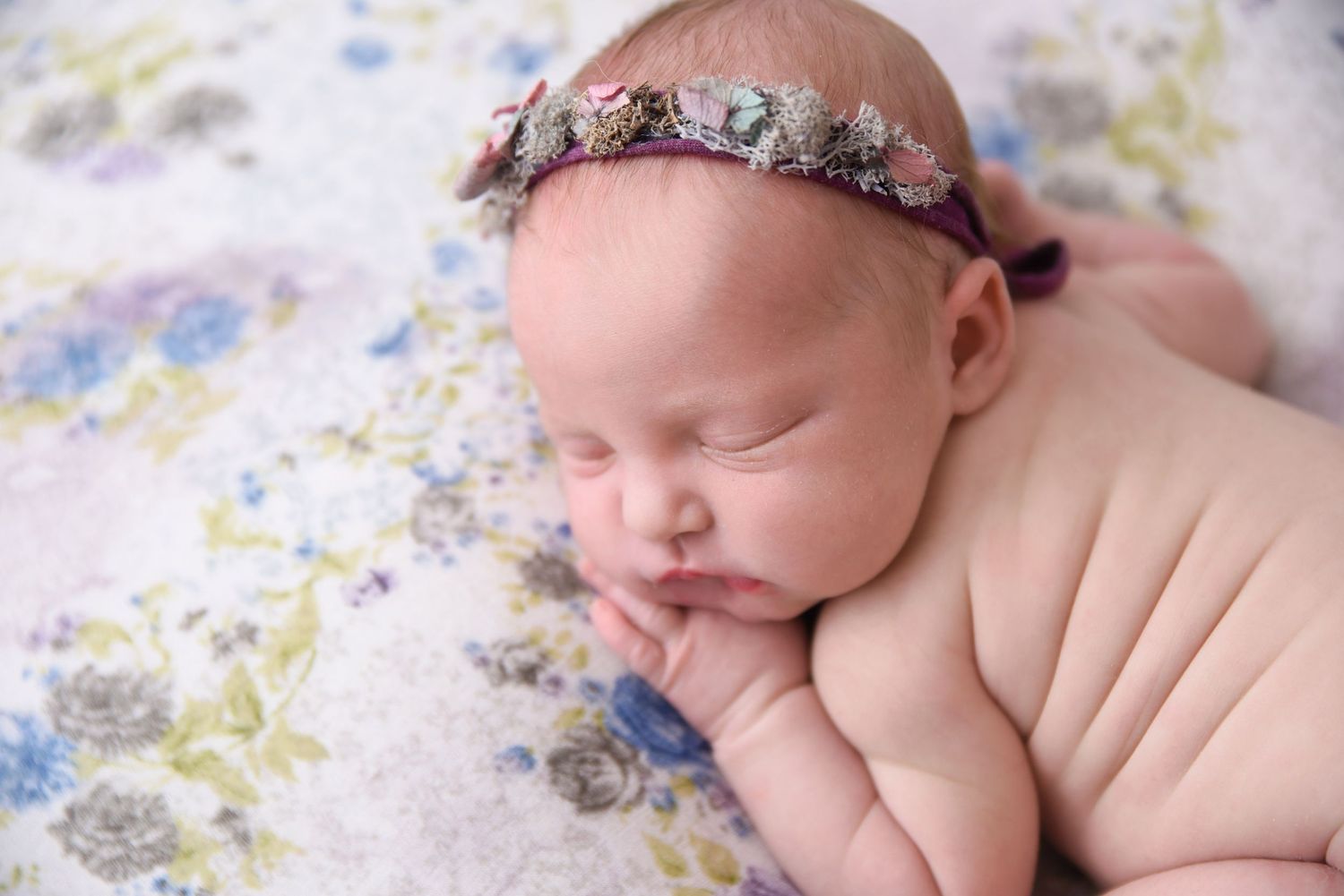 Peaceful newborn baby girl wearing floral headband sleeping on soft floral blanket in professional portrait.
