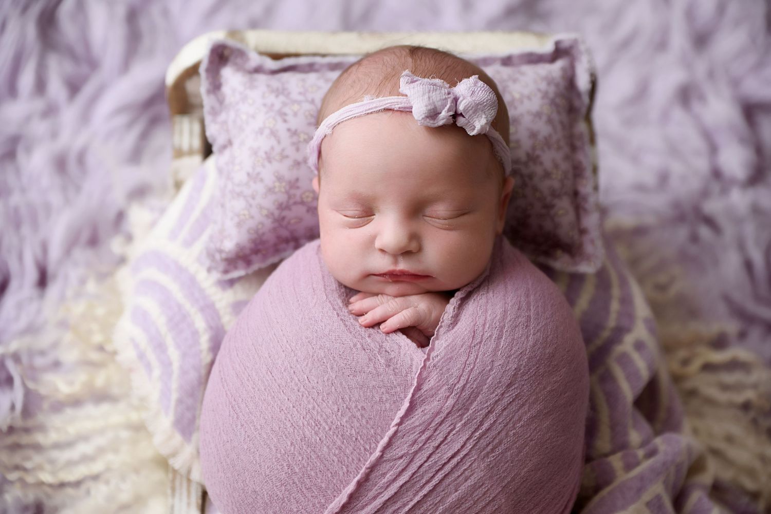 Newborn baby girl swaddled in pink wrap with bow headband sleeping in tiny bed during studio session.