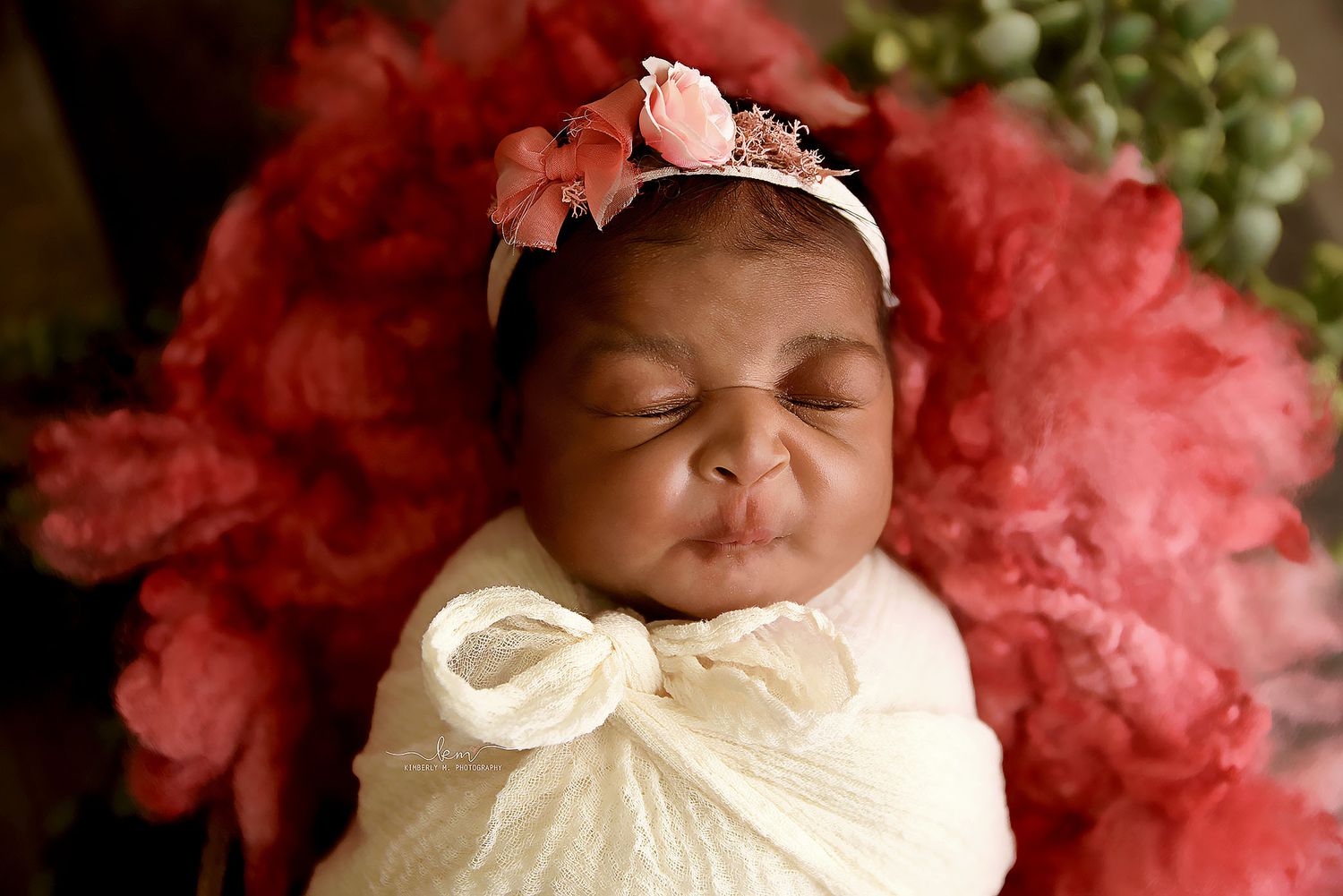 Newborn baby girl swaddled in white wrap with pink floral headband resting on a bed of coral flowers.