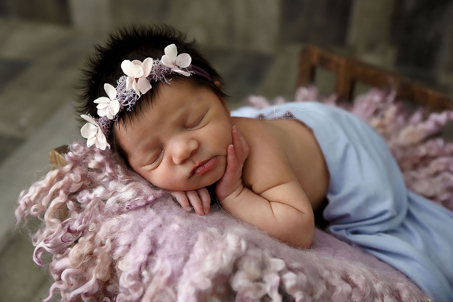 Sleeping newborn girl with black hair wearing a floral headband on purple fur.