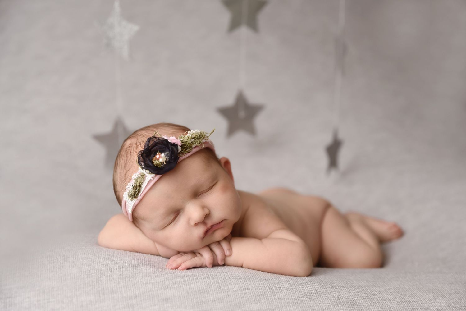 Sleeping newborn girl wearing a floral headband lying on a gray blanket with star decorations above.