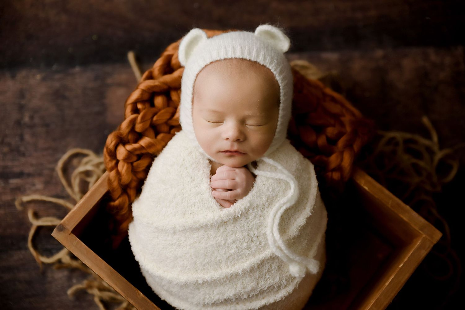 Newborn baby wrapped in cream swaddle wearing bear bonnet sleeping in wooden box during studio portrait.