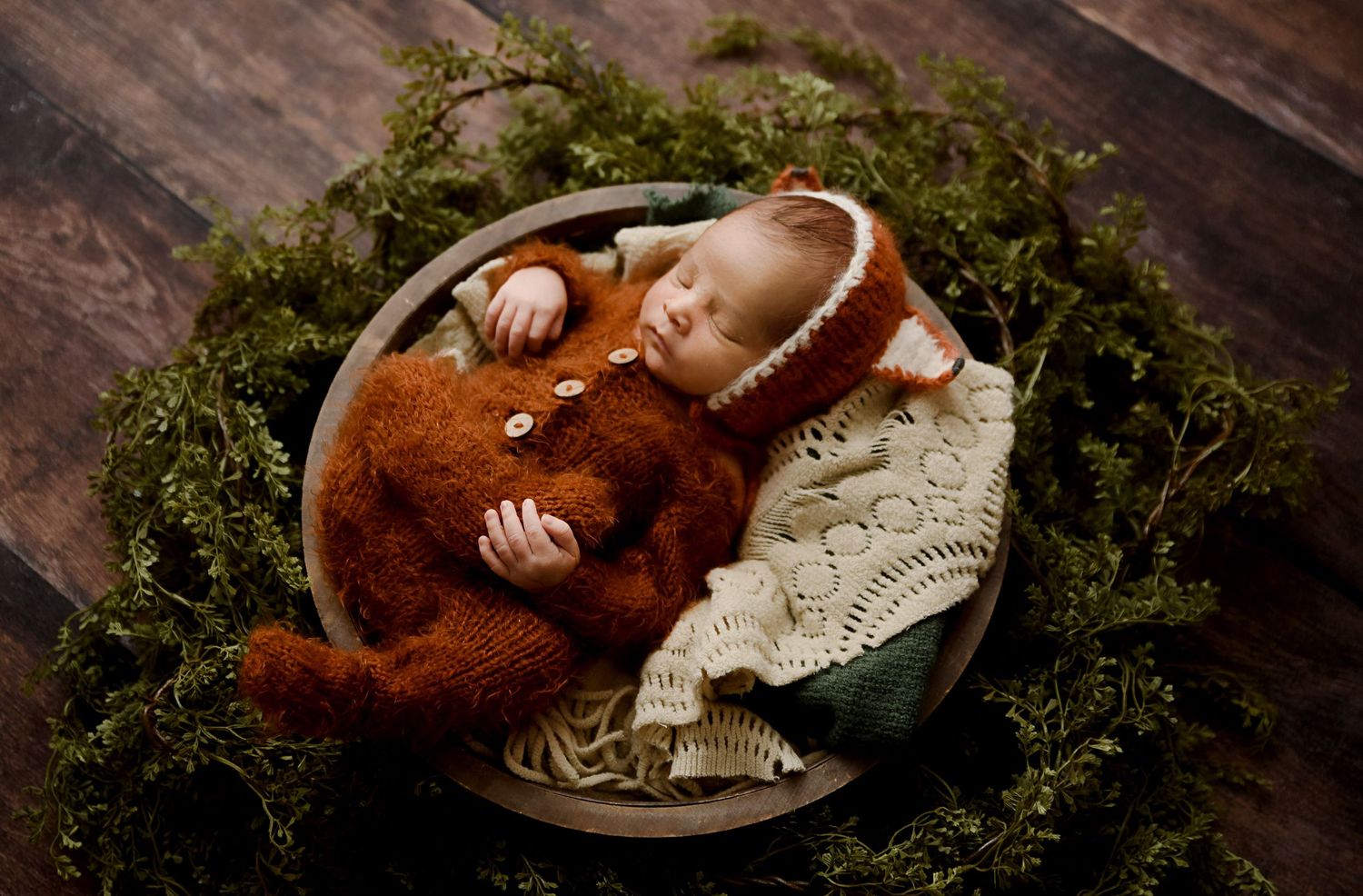 Sleeping newborn baby in rust knit outfit cuddling teddy bear in wooden bowl with green moss backdrop.