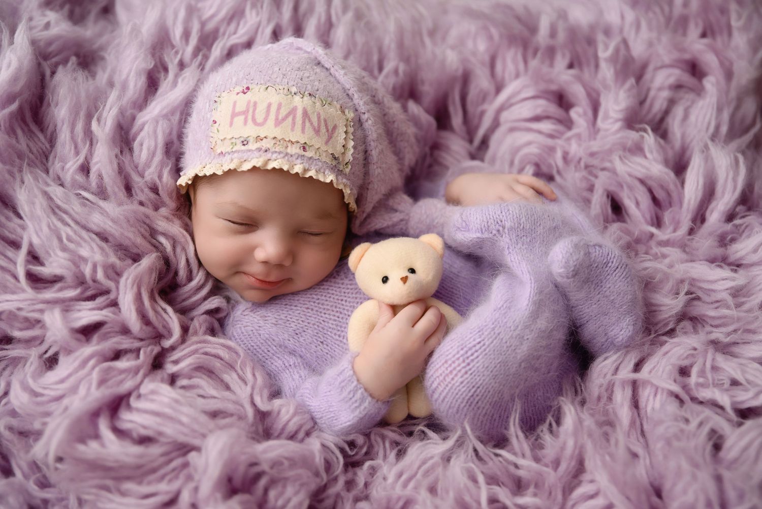 Sleeping newborn in purple outfit holding a teddy bear wearing a knit hat on a lavender fur blanket.