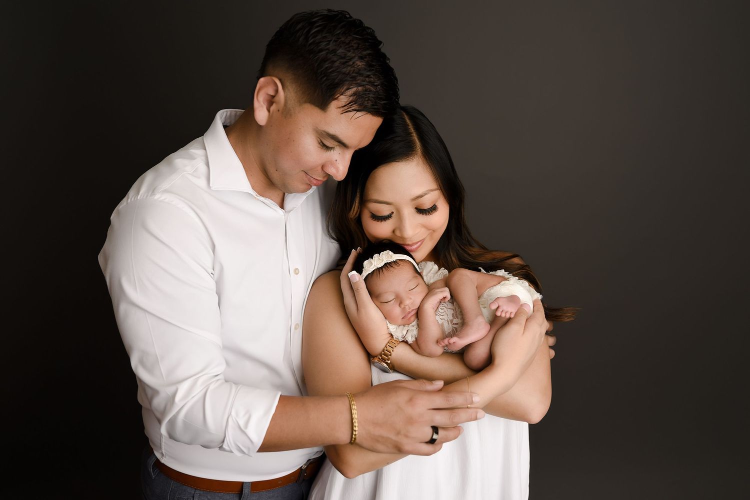 Parents in white clothing tenderly holding their sleeping newborn baby against a dark gray background.
