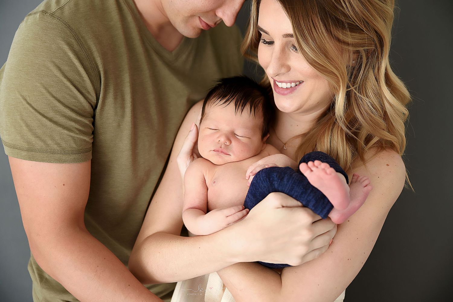 Smiling parents lovingly gazing at their sleeping newborn baby wrapped in navy blue in a studio portrait.