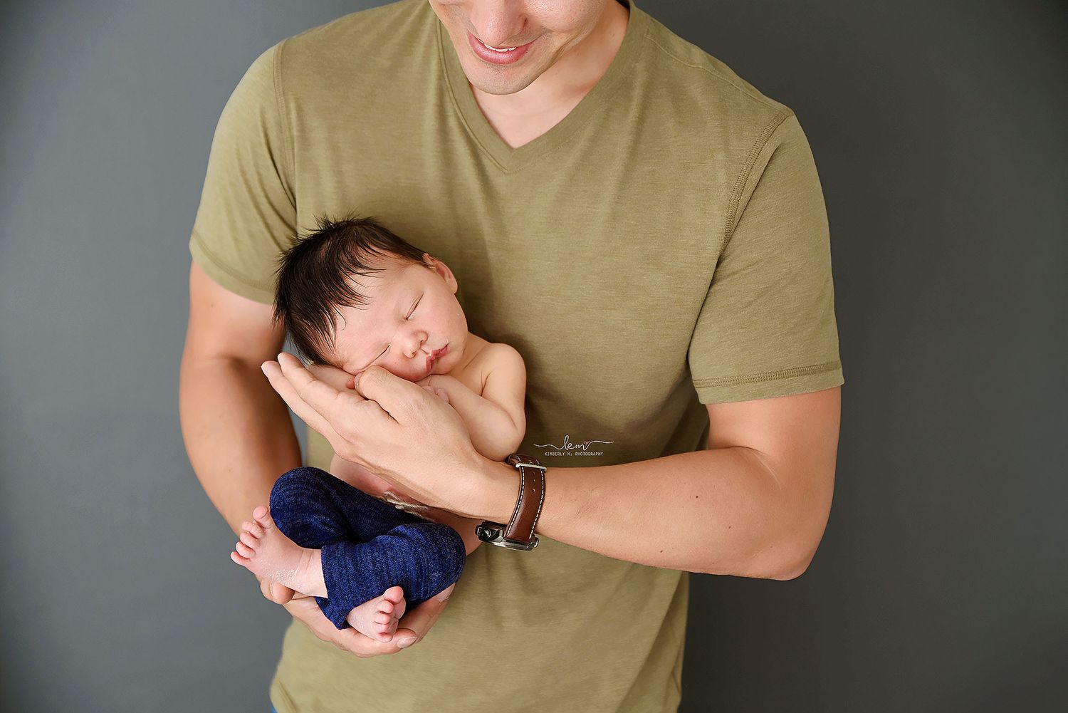 Father in olive green shirt cradling sleeping newborn baby wrapped in navy blue against a gray background.