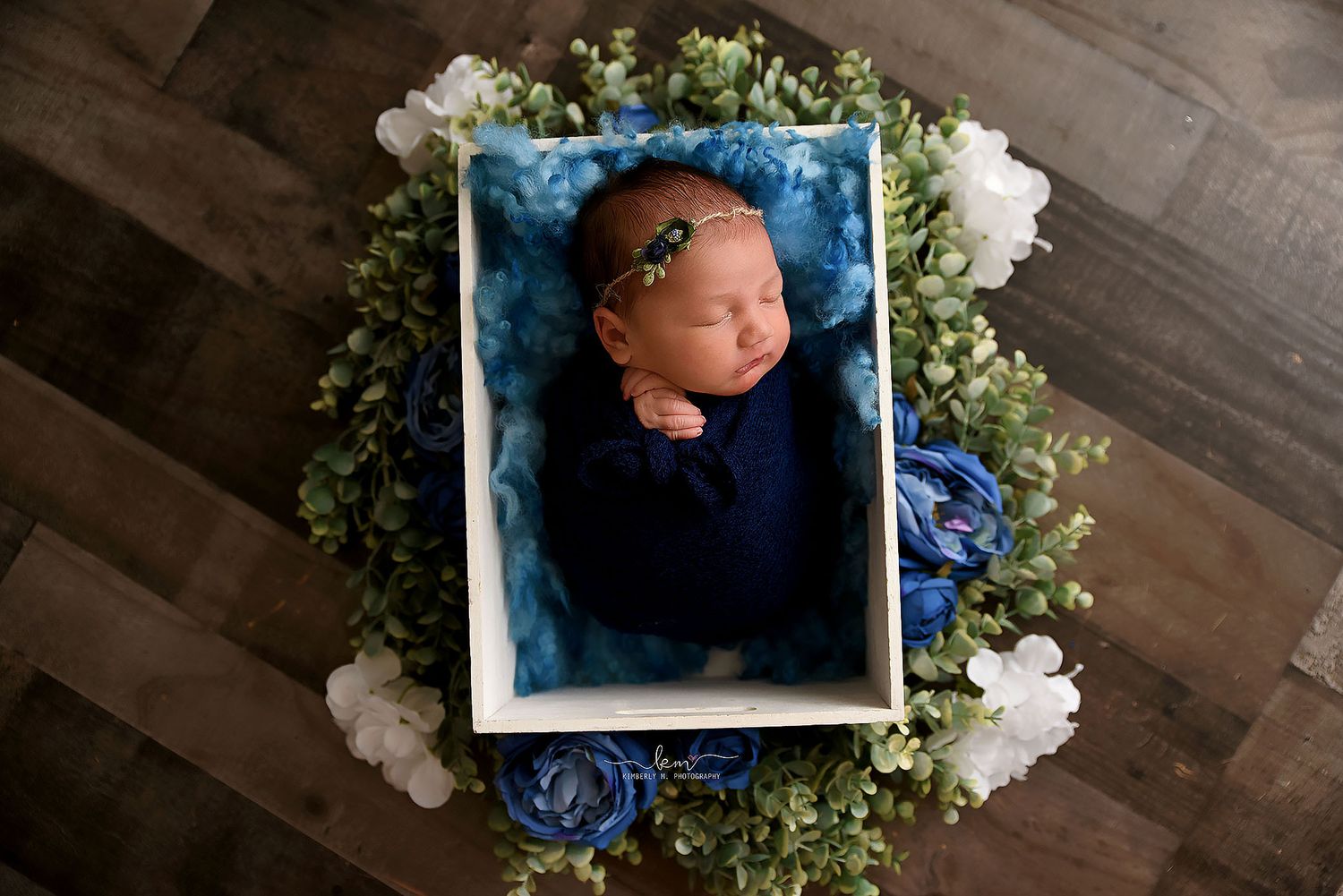 Newborn baby swaddled in navy blue wrap lying in a white box surrounded by green foliage and blue flowers.